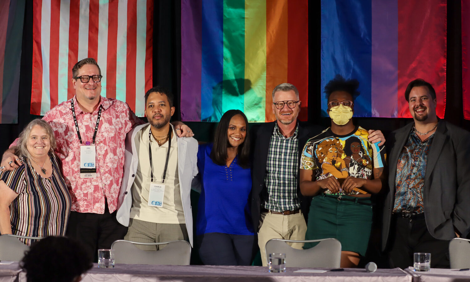 Seven smiling panelists from an NLGJA convention session standing close together on a stage behind a desk against a background of American, Pride, and Bi+ flags.